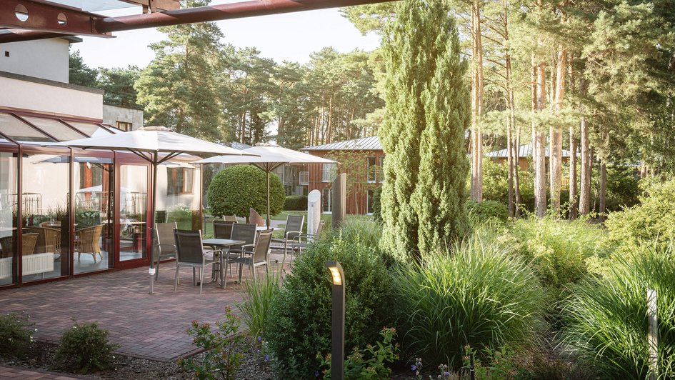 Terrasse mit Sitzgelegenheiten unter einem Sonnenschirm und Sträuchen und Bäumen drumherum. 