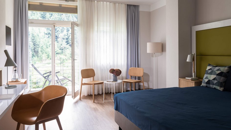 Patientenzimmer mit großem Bett, Sitzgelegenheiten und offener Tür zum Balkon