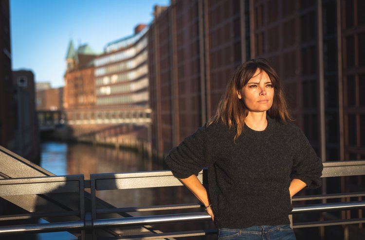 Frau mit schwarzem Pullover und blauer Jeans steht auf einer Brücke und lehnt sich ans Geländer